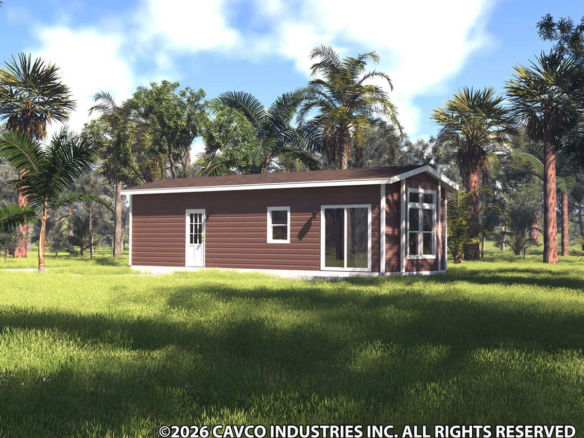 A small brown manufactured home with white trim stands in a grassy clearing surrounded by trees under a partly cloudy sky.