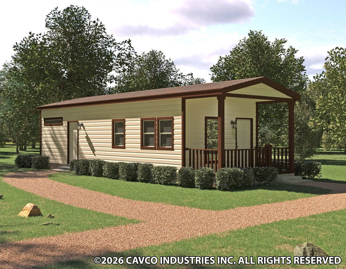 A single-story manufactured home with a covered porch, cream siding, and brown trim, surrounded by trees and grass, with a gravel walkway in front.