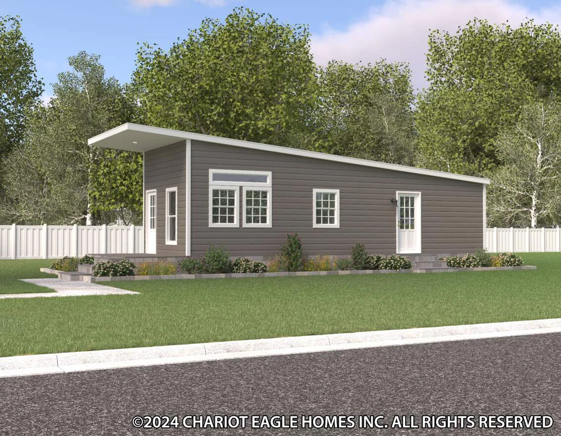 A modern, single-story gray home with a slanted roof sits on a green lawn, surrounded by trees and a white fence.