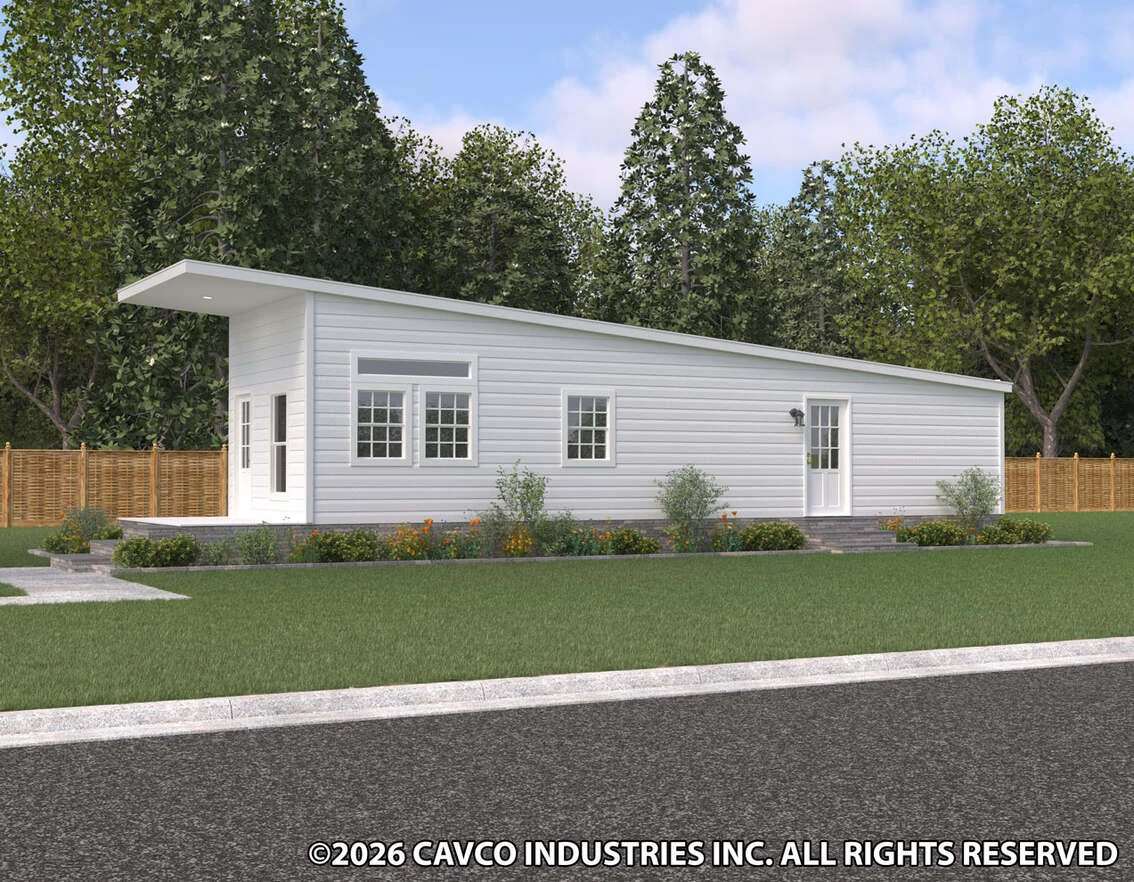 Single-story modern manufactured home with white siding, large windows, and a sloped roof, surrounded by grass, shrubs, and trees, with a wooden fence in the background.