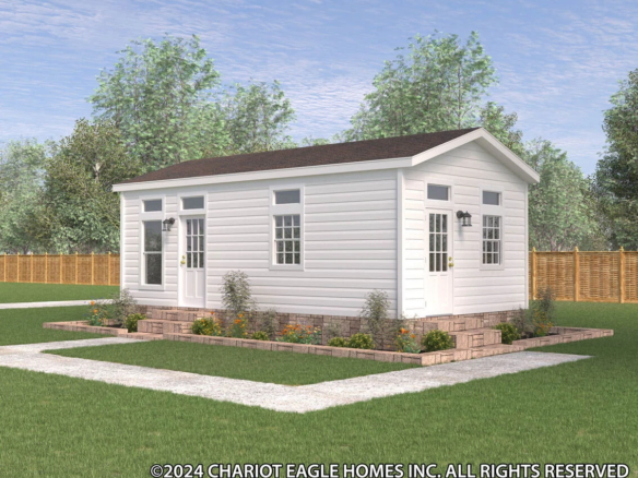 A small white modular home with a gable roof, double doors, multiple windows, and a landscaped garden, set in a fenced yard with trees in the background.
