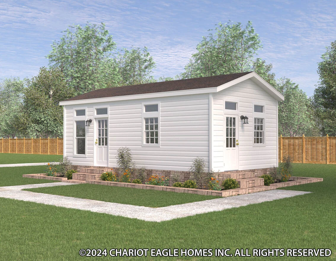 A small white modular home with a gable roof, double doors, multiple windows, and a landscaped garden, set in a fenced yard with trees in the background.