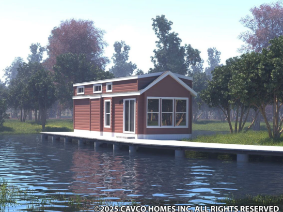 A small red house with white trim sits on a dock over a calm lake, surrounded by trees and grassy areas under a bright sky.