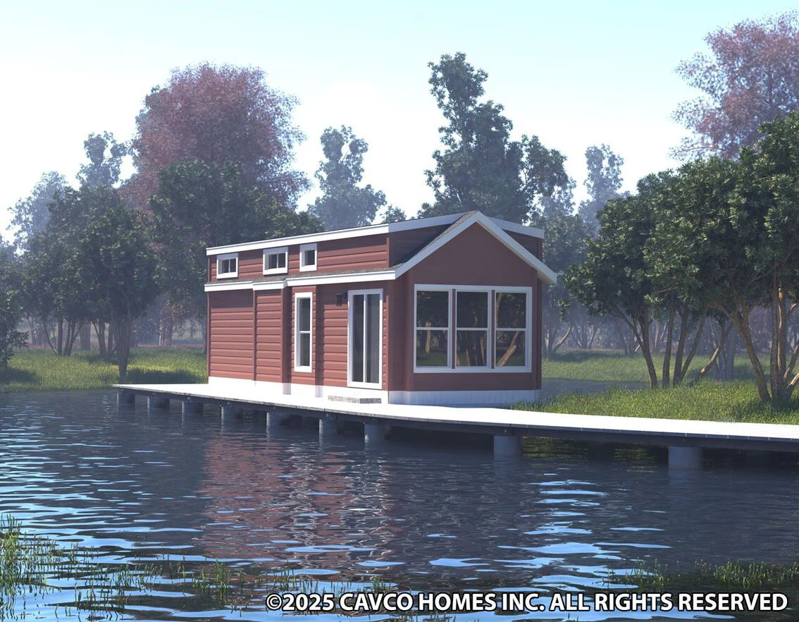 A small red house with white trim sits on a dock over a calm lake, surrounded by trees and grassy areas under a bright sky.