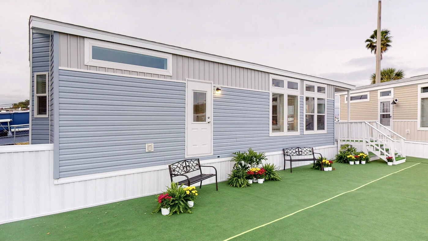 A light blue and white mobile home with large windows, a central door, and two black benches with potted plants arranged on artificial green turf in front.