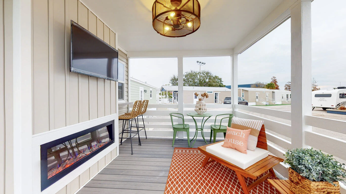A screened-in porch with a mounted TV, electric fireplace, two barstools, a small green table with chairs, decorative pillows, and an orange patterned rug.