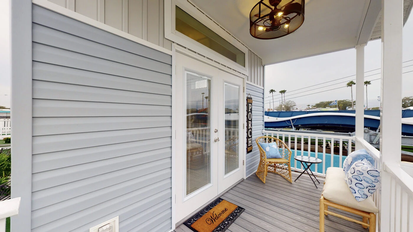 A small front porch with light gray siding, a Welcome doormat, glass double doors, two wicker chairs with cushions, a side table, and a decorative hanging fan above.