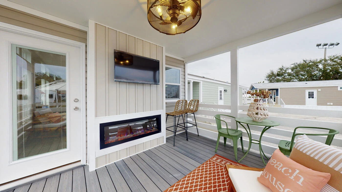Covered porch with wood flooring, wall-mounted TV, electric fireplace, bar stools at a counter, green chairs with a table, and decorative pillows on a sofa.