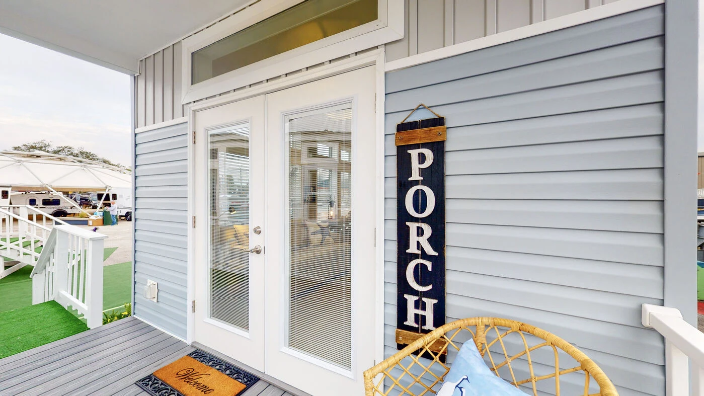 Glass double doors on a light blue house exterior, with a vertical sign reading "PORCH," a welcome mat, and part of a wicker chair visible in the corner.