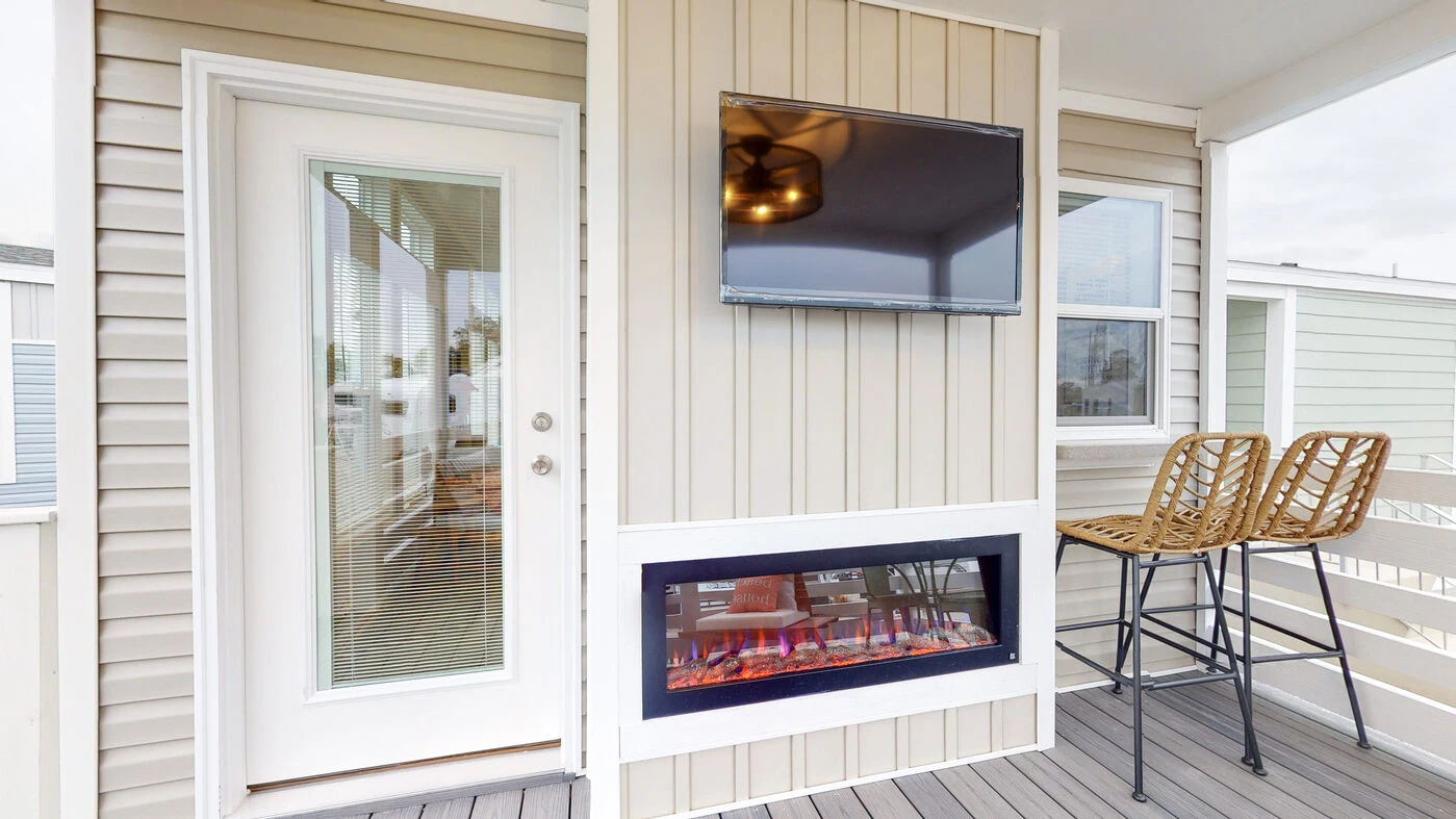 A small outdoor patio features a wall-mounted TV, an electric fireplace, two wicker bar stools, and a glass door with blinds leading inside.