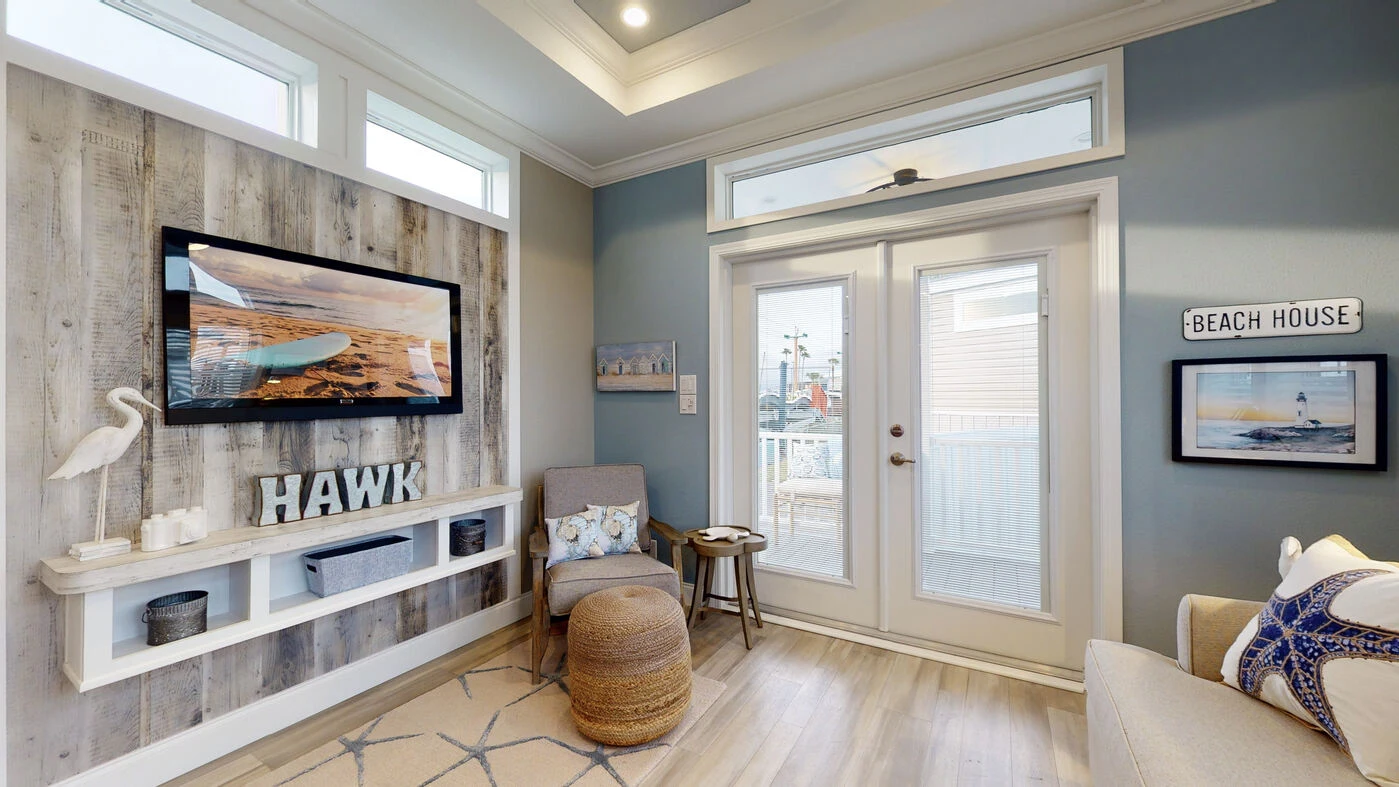 Small coastal-themed living room with light wood floors, a mounted TV, beach decor, armchair, ottoman, and double glass doors leading outside.