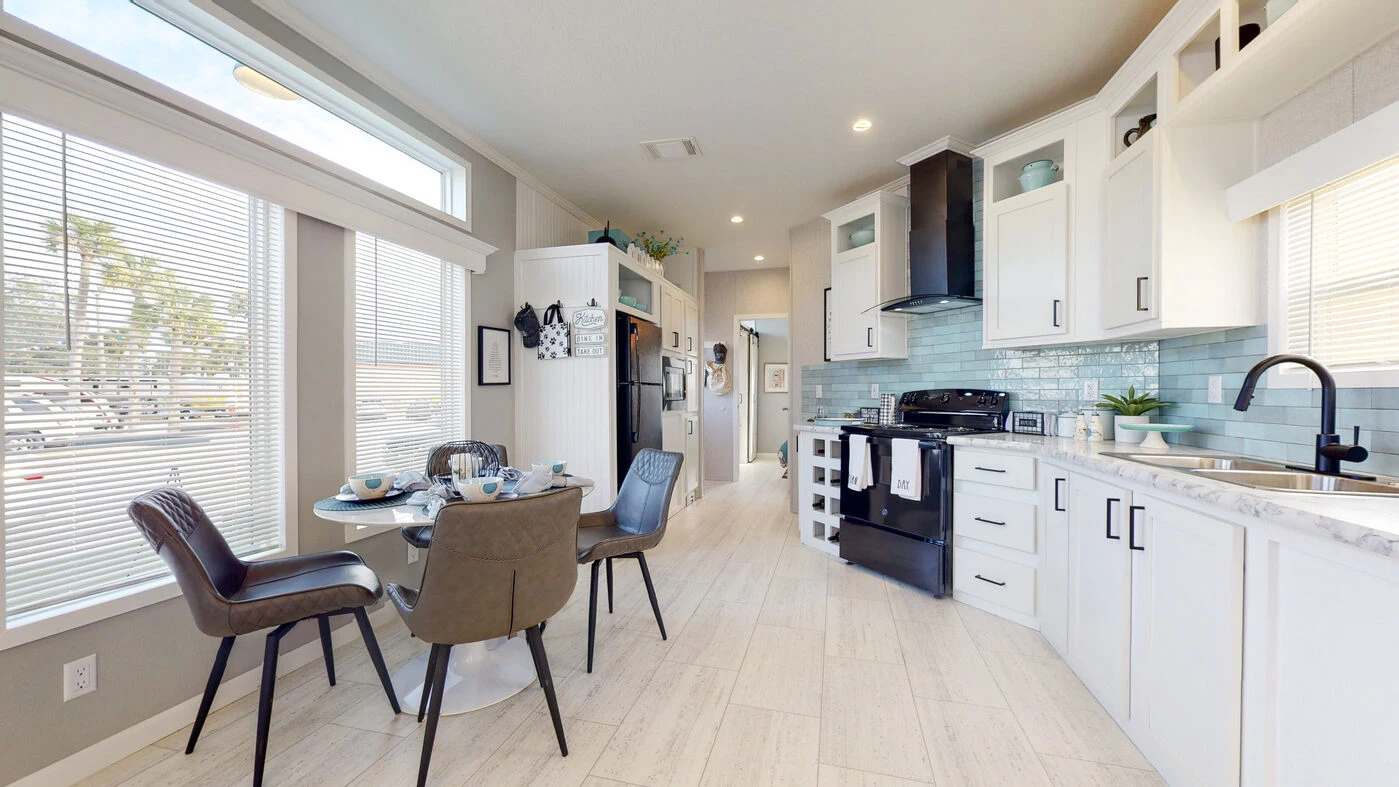 Modern kitchen with white cabinets, black appliances, a round dining table set for four, large windows, light wood flooring, and blue tile backsplash.