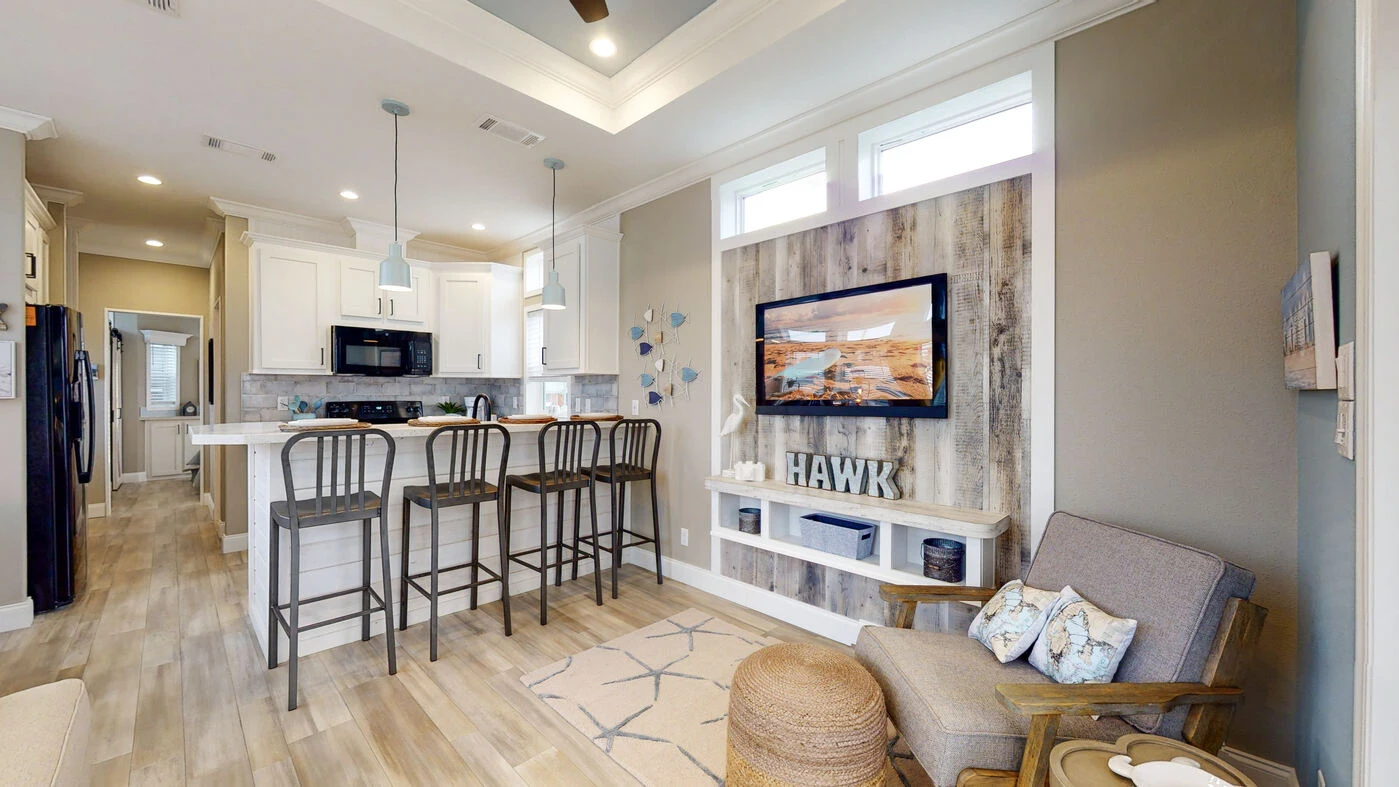 Open-concept living area with a kitchen, three bar stools at the counter, a wall-mounted TV, and neutral-toned decor.