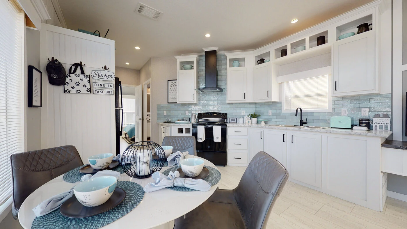 Modern kitchen with white cabinets, blue tile backsplash, black appliances, light blue accents, and a round dining table set for four with blue placemats and dishware.