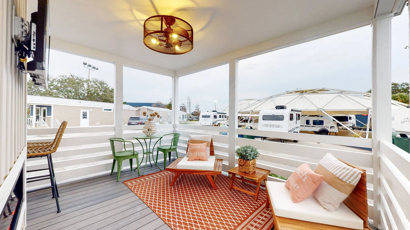 Covered porch with white railing, two cushioned wooden benches, a small coffee table, green bistro set, potted plants, and a geometric rug. Dome structure and trailers visible outside.