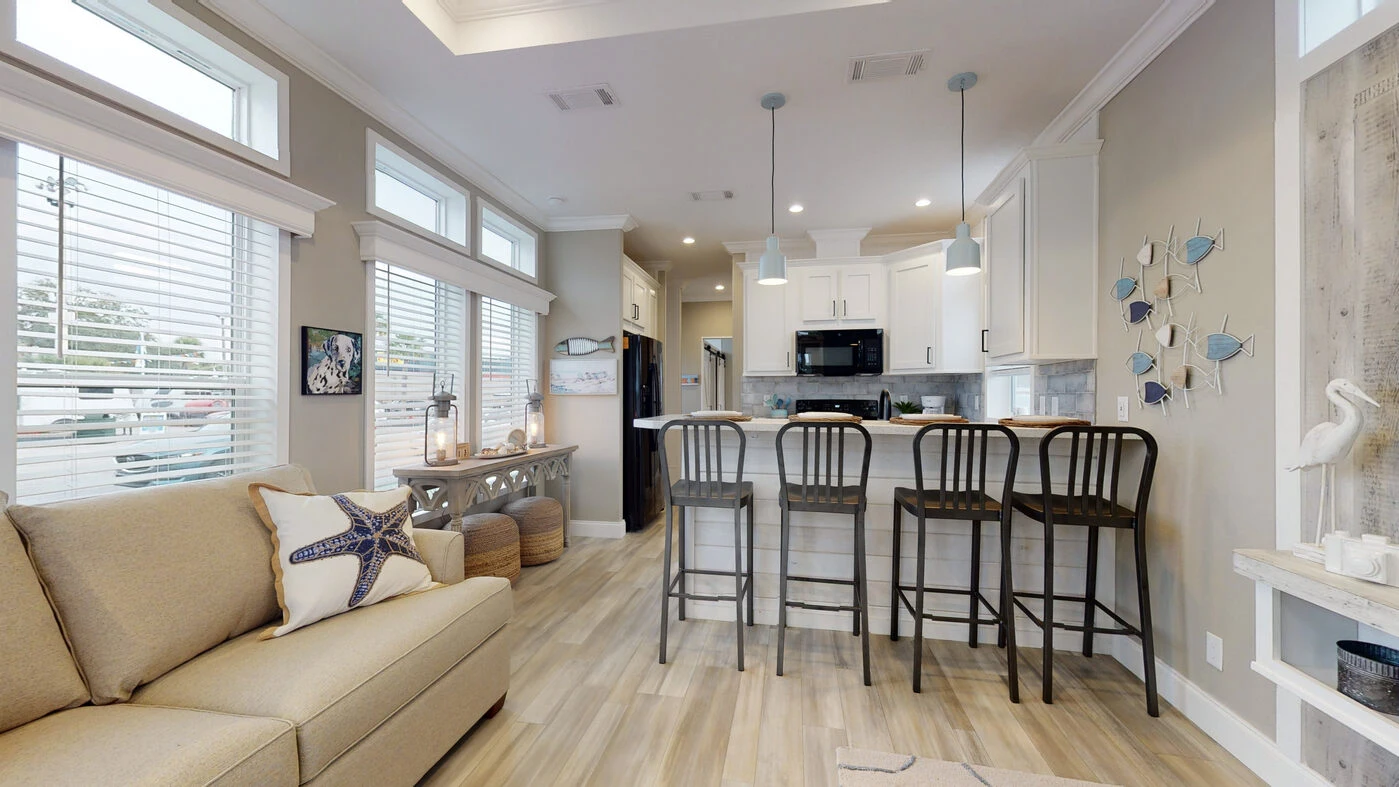 Modern kitchen and living area with light wood floors, beige sofa, breakfast bar with three chairs, white cabinets, and nautical-themed decor.