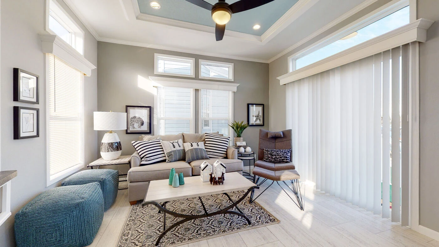 Modern living room with a gray sofa, patterned pillows, armchair, ottomans, coffee table, large windows, and vertical blinds letting in natural light.