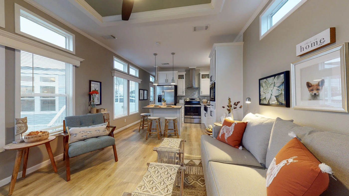 Modern living room and kitchen with neutral colors, wooden flooring, light gray walls, cozy furniture, and decor including framed art and throw pillows.