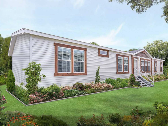 A white manufactured home with brown trim and multiple windows sits on a well-maintained lawn, surrounded by flowering shrubs and greenery.
