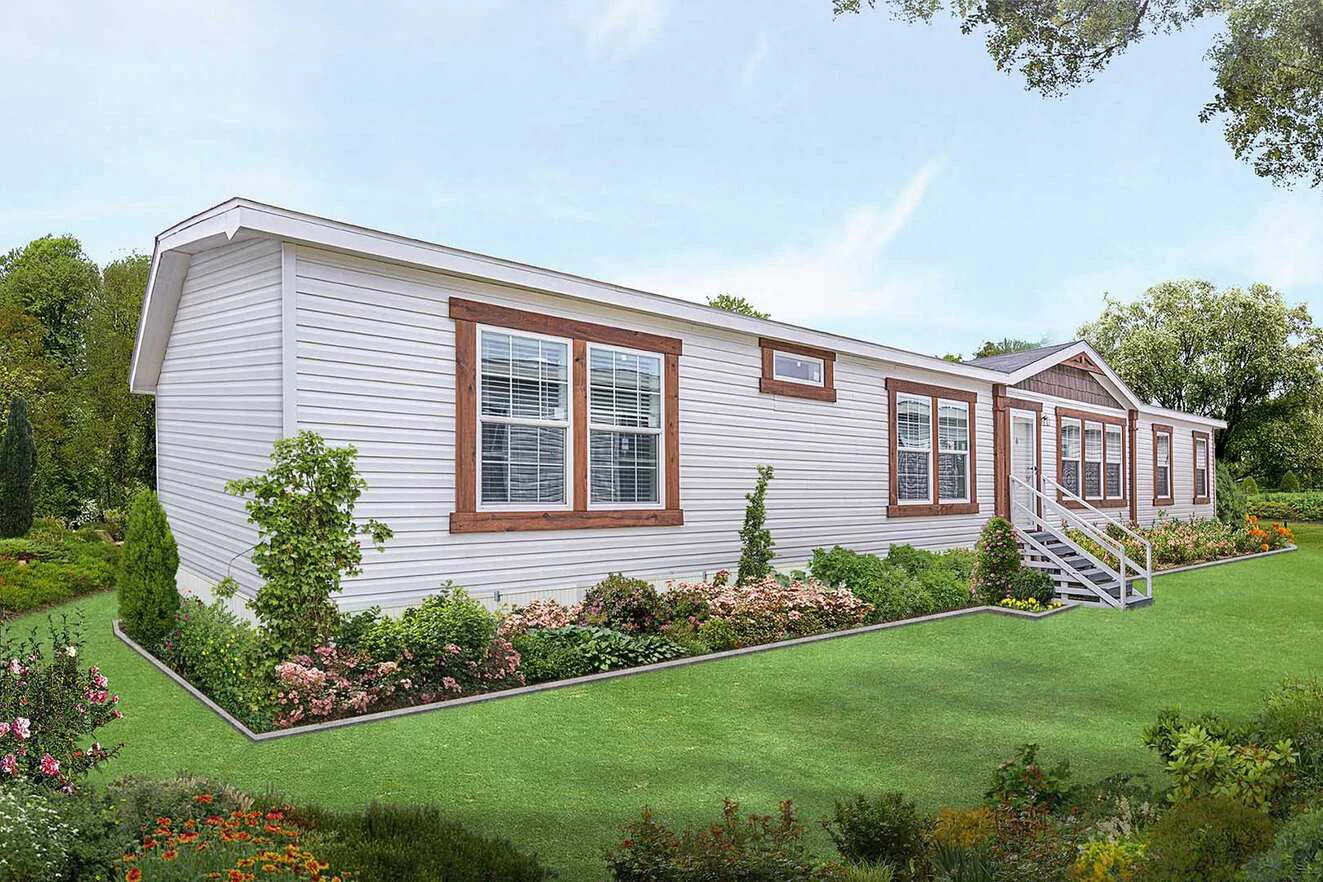 A white manufactured home with brown trim and multiple windows sits on a well-maintained lawn, surrounded by flowering shrubs and greenery.