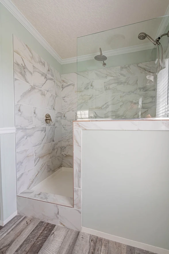 Modern walk-in shower with marble-patterned wall tiles, clear glass partition, overhead showerhead, and wood-look flooring outside.