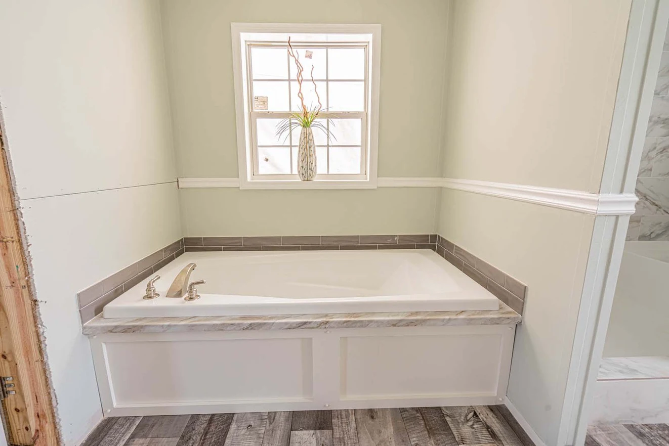 White built-in bathtub with gray tile trim under a window, featuring a plant in a vase on the windowsill; partial view of wood flooring and unfinished door frame.