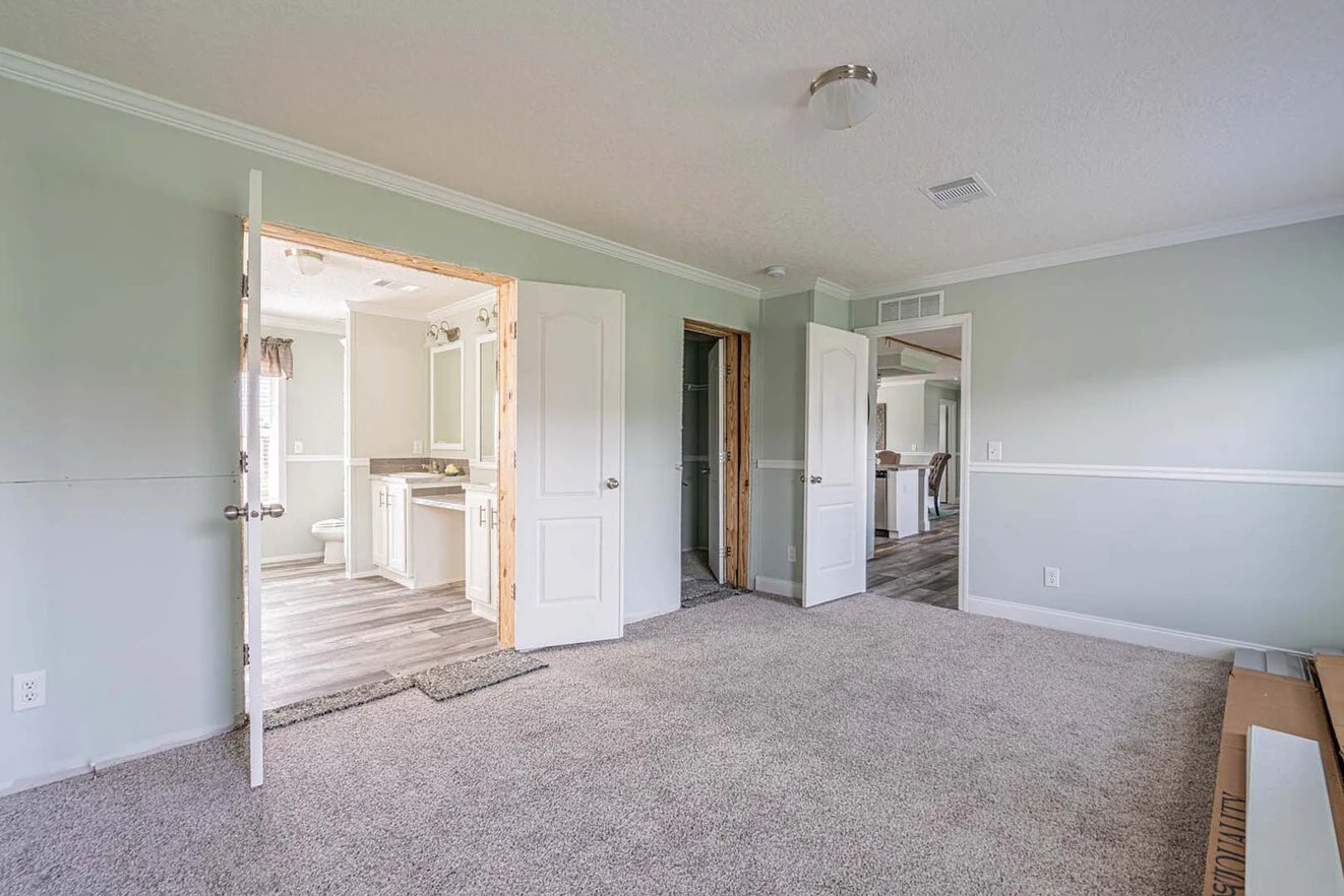 Empty carpeted bedroom with light green walls, two open doors leading to a bathroom and a closet, and a window in the background. Boxes are stacked on the right side of the room.