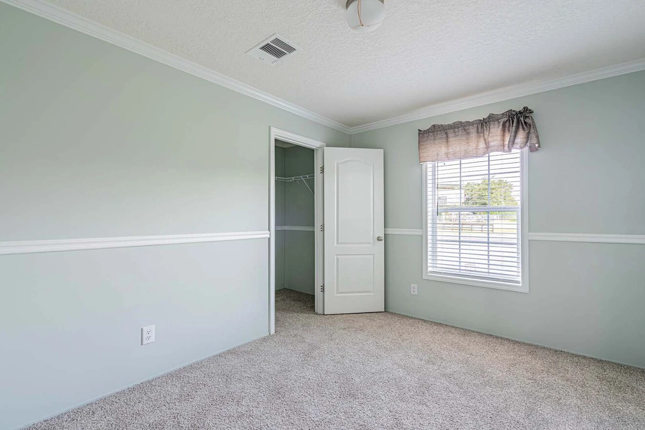 Empty bedroom with light gray walls, carpeted floor, open white door leading to a closet, window with blinds and a valance, and a ceiling light fixture.