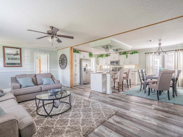 Open-concept living and dining area with light gray sofas, glass coffee table, dining set, kitchen island, ceiling fans, and wood-look flooring. Greenery decor tops white cabinets.