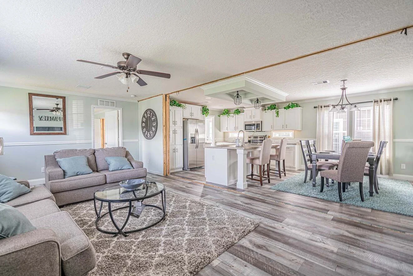 Open-concept living and dining area with light gray sofas, glass coffee table, dining set, kitchen island, ceiling fans, and wood-look flooring. Greenery decor tops white cabinets.