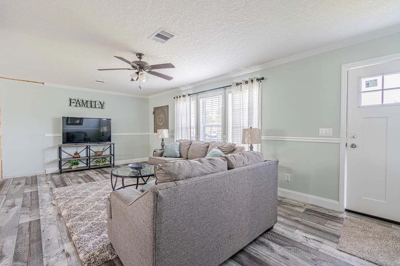 Bright living room with gray sectional sofa, round glass coffee table, TV on a stand, large windows with curtains, and light wood flooring.