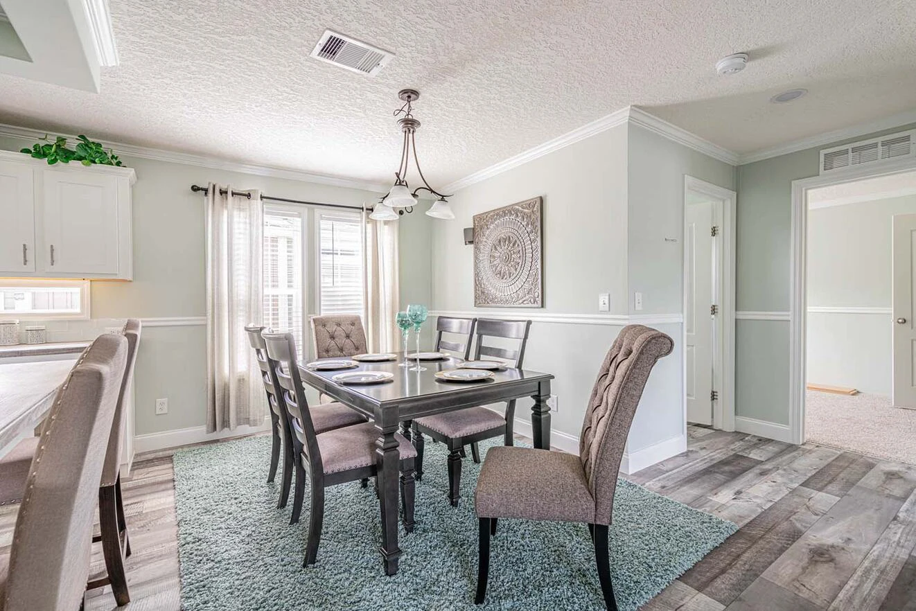 A dining room with a dark wood table set for six, upholstered chairs, a light blue rug, and a large decorative wall art piece; natural light coming through two windows.