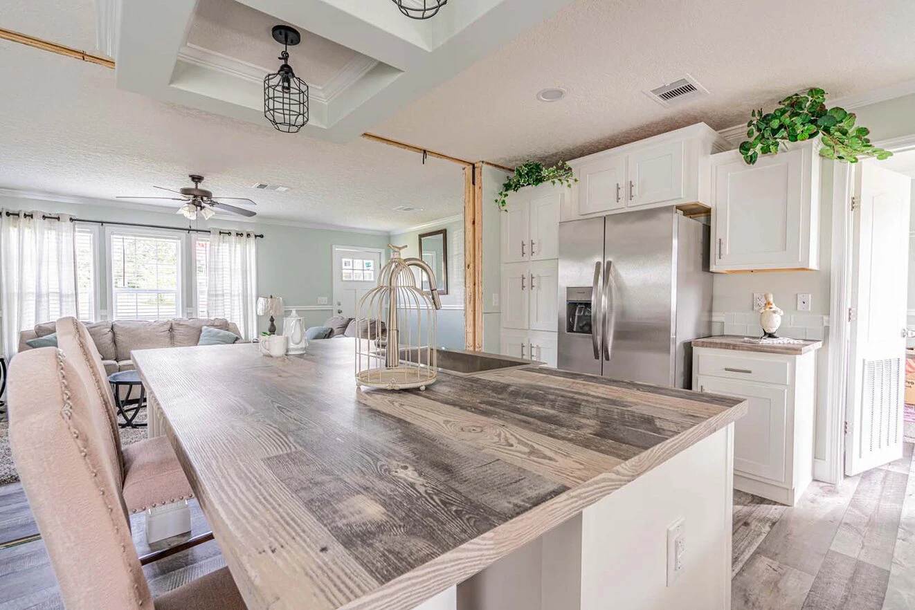 Bright kitchen with a wooden island, barstools, stainless steel refrigerator, white cabinets, and a view into a living room with a sectional sofa and large windows.