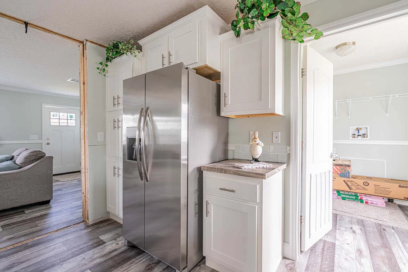 A modern kitchen with white cabinets, a stainless steel refrigerator, potted plants on top, and boxes stacked in an adjacent room.