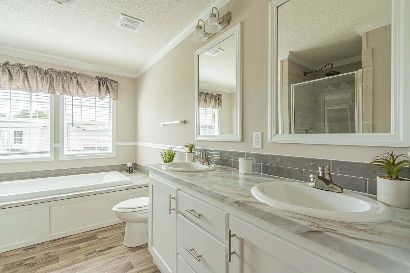 Bright bathroom with a double sink vanity, marble countertop, large mirror, bathtub by the window, toilet, and light wood flooring.