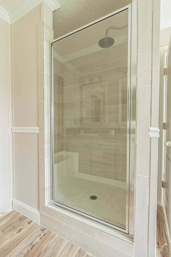 Glass-enclosed shower with a rainfall showerhead, beige tile walls, and wood-patterned vinyl flooring outside the shower.