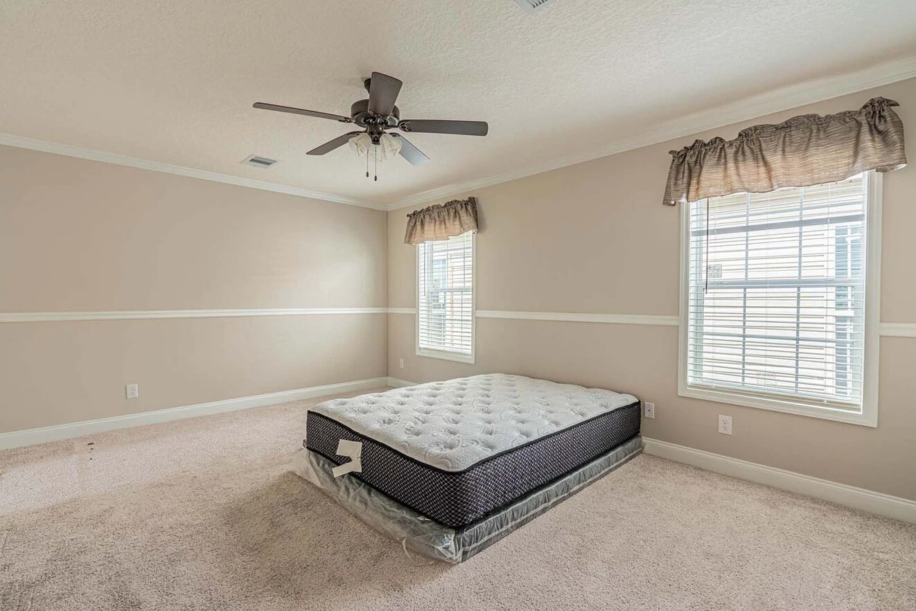 A sparsely furnished bedroom with beige walls, two windows with blinds and valances, a ceiling fan, and a bare mattress on the carpeted floor.