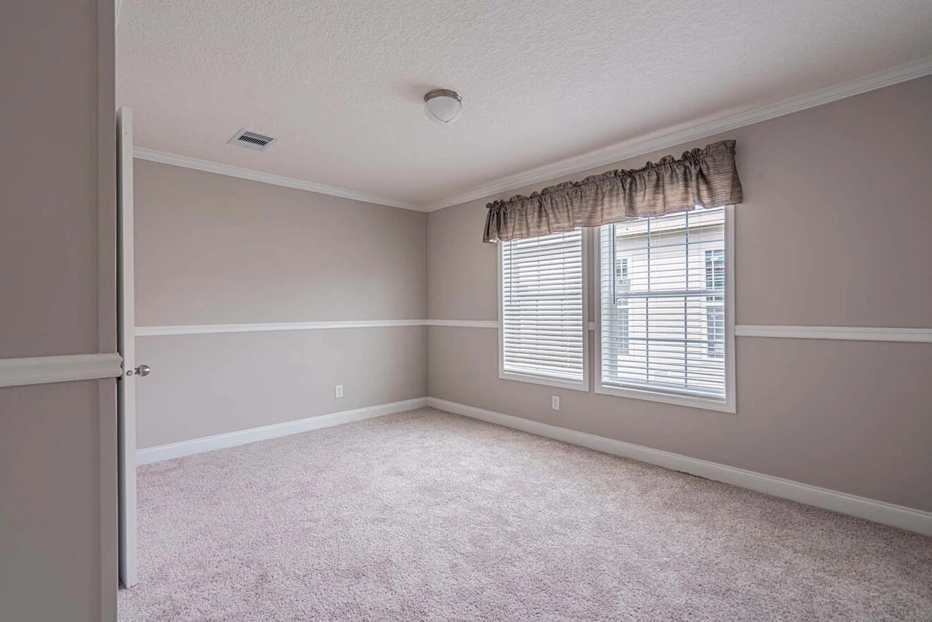 Empty carpeted room with beige walls, white trim, a ceiling light, two windows with blinds and a valance, and an open door on the left.
