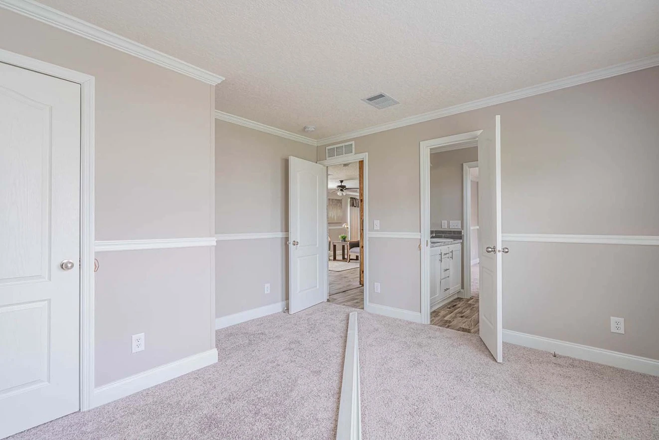 Empty bedroom with beige walls, white trim, and carpeted floor; doors open to adjacent hallway and bathroom.