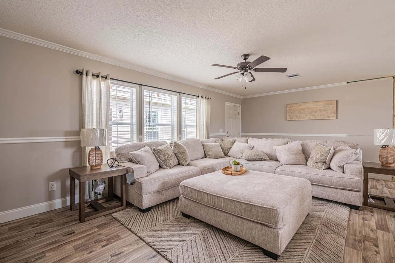 A neutral-toned living room with a large sectional sofa, ottoman, wooden side tables, lamps, and windows with white curtains letting in natural light.