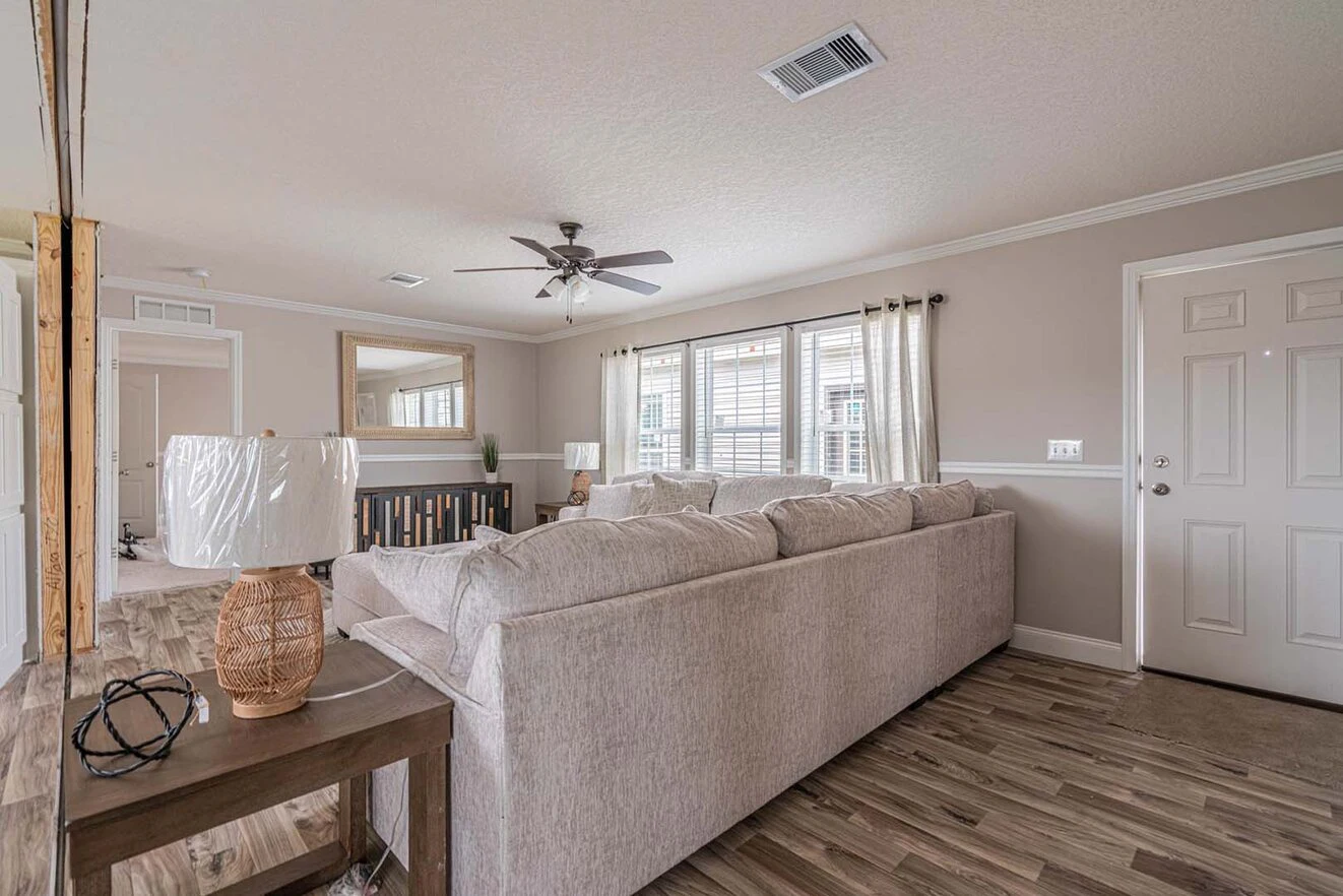 Neutral-toned living room with a large sectional sofa, side table, lamps, ceiling fan, wood flooring, and windows with curtains letting in natural light.