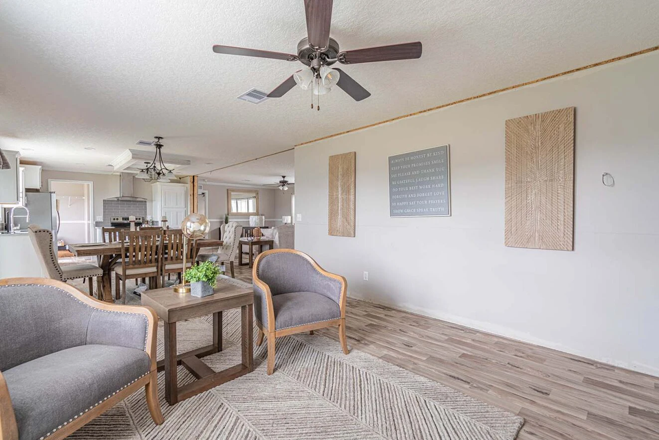 A bright living and dining area with wood flooring, two gray armchairs, a small table, dining set, ceiling fans, wall art, and neutral decor.