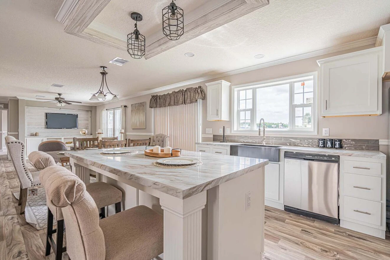 A modern kitchen with white cabinets, marble island, stainless steel appliances, and beige upholstered chairs, connected to a living area with a TV and wood flooring.