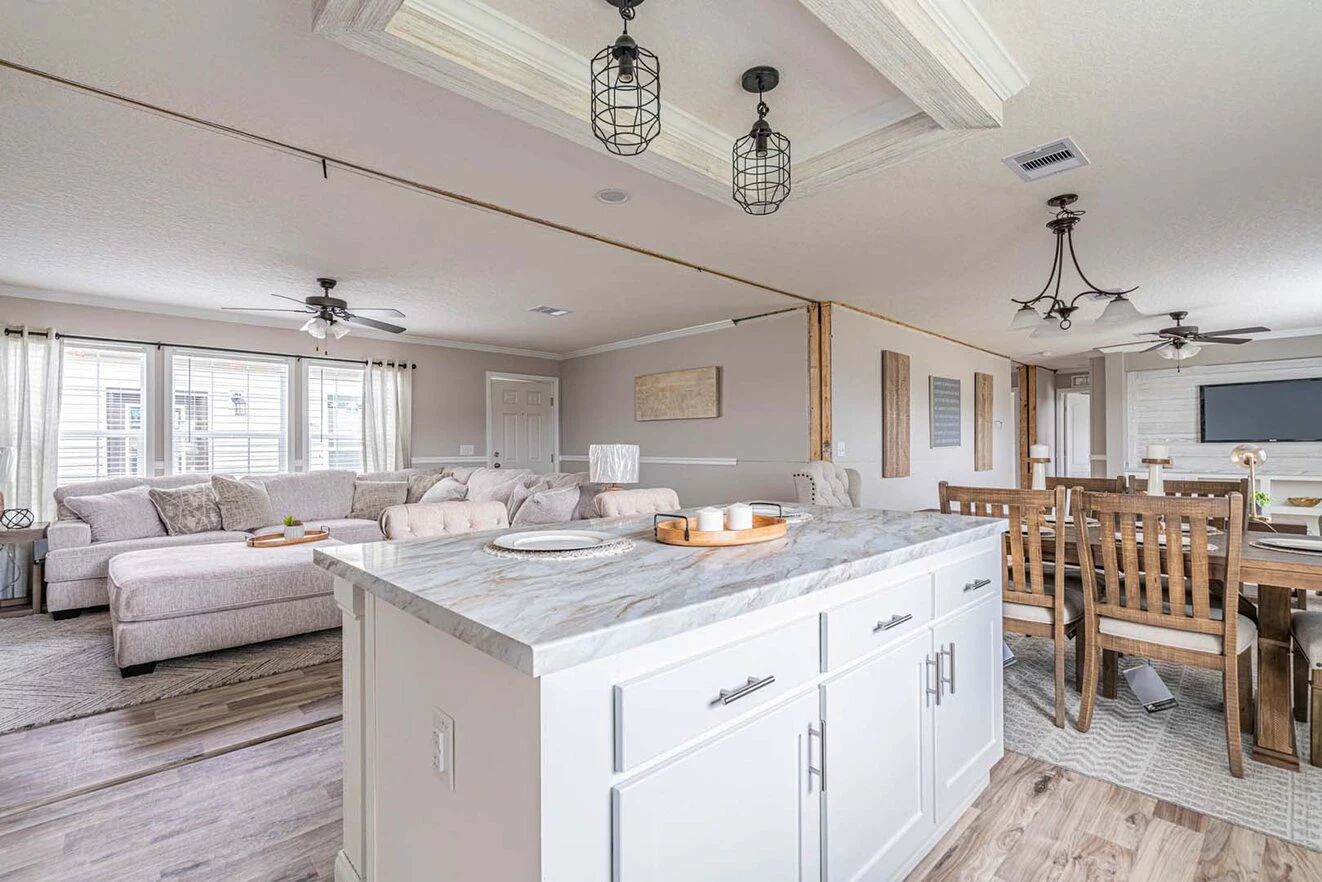 Open-concept living and dining area with a marble kitchen island, white cabinets, wooden dining table, and a large sectional sofa by windows.