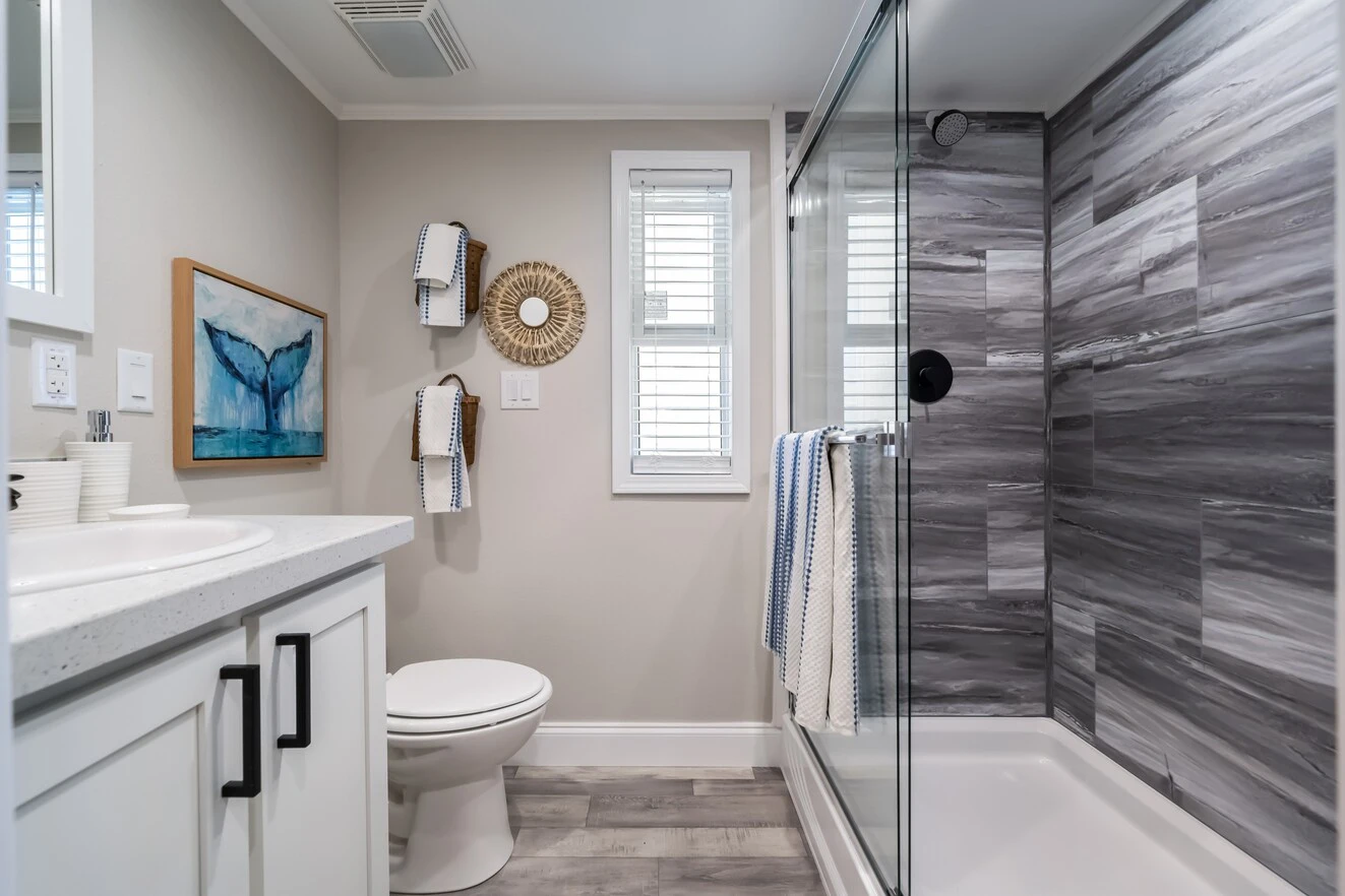 chariot_eagle_eagle_eg12341a_bath_0598_1_1673903478285_1323_12 Modern bathroom with a glass-enclosed shower, gray and white tile, a toilet, a white vanity, wall art, towel racks, and a window with white blinds.