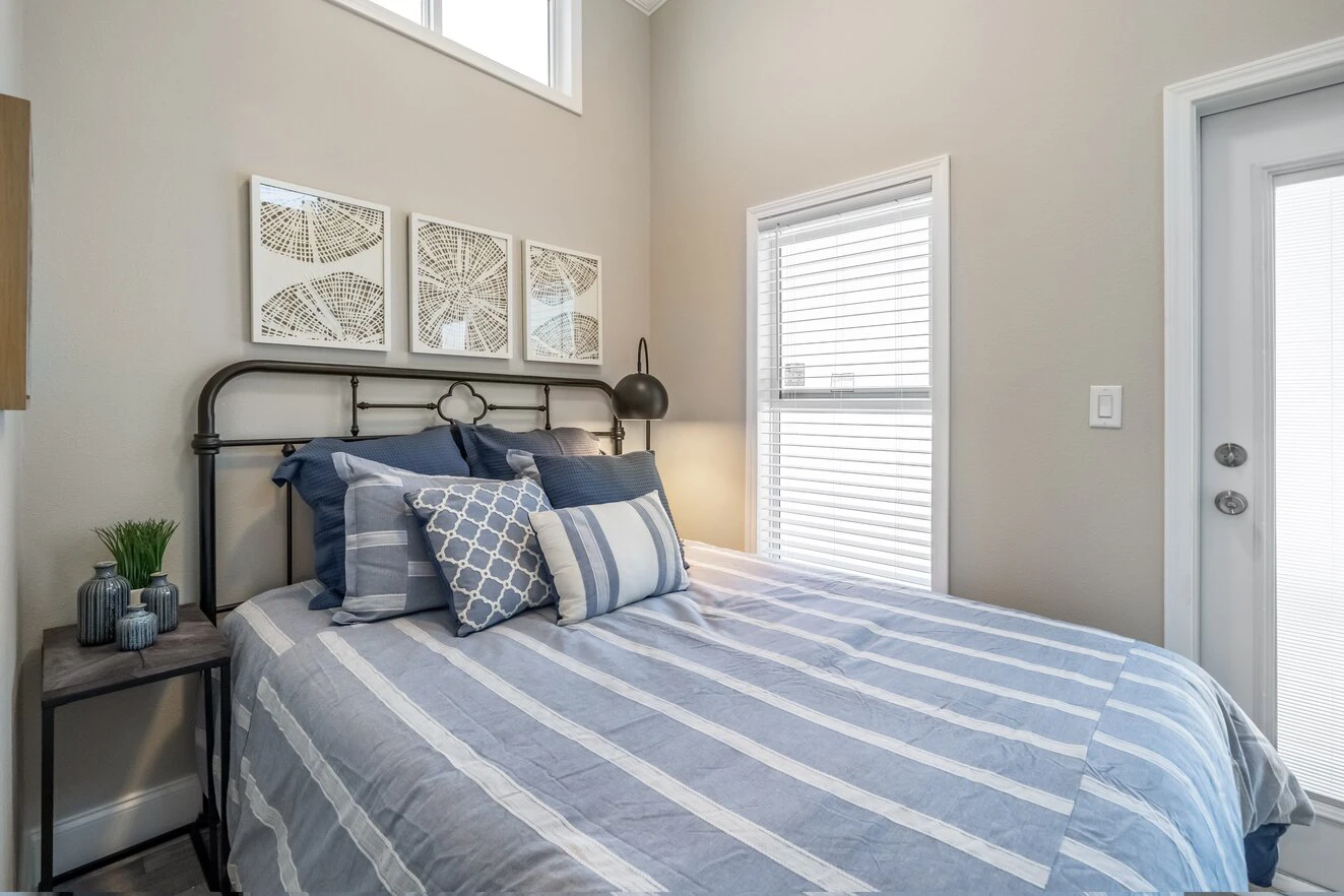 chariot_eagle_eagle_eg12341a_bedroom_0711_1_1673903459166_1323_12 A neatly made bed with blue and white striped bedding, decorative pillows, and a metal headboard sits by a window with blinds in a light-colored bedroom.