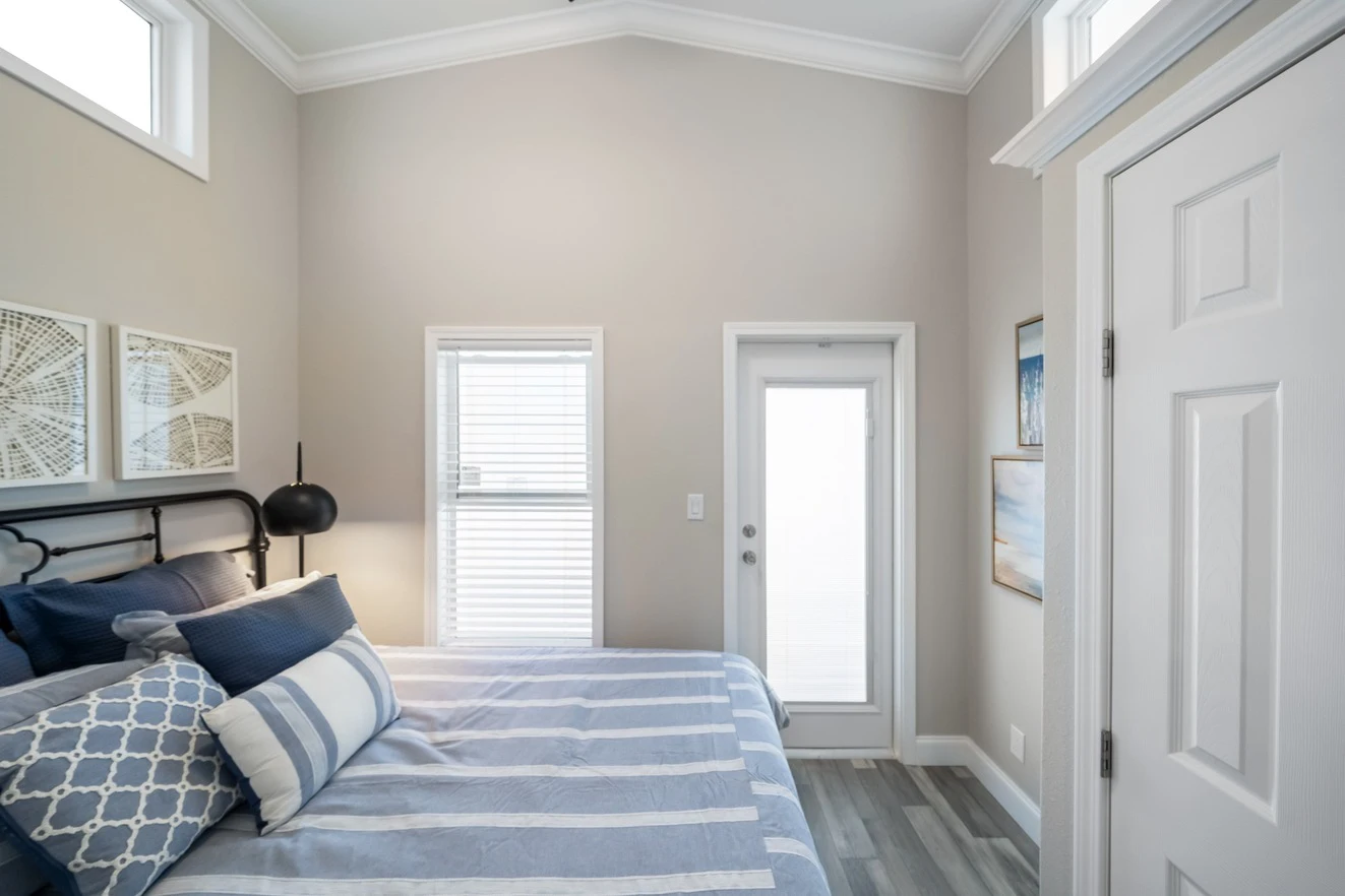 chariot_eagle_eagle_eg12341a_bedroom_0849_1_1673903446362_1323_12 A small, modern bedroom with a striped bed, neutral walls, large windows, light wood flooring, and framed artwork on the walls.