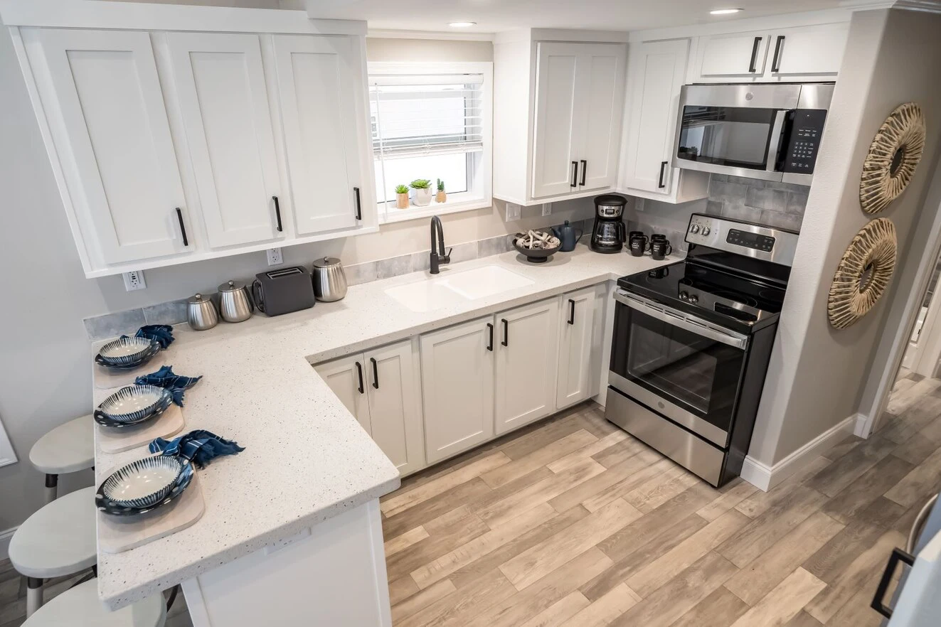 chariot_eagle_eagle_eg12341a_kit_0348_1_1673903428511_1323_12 Modern kitchen with white cabinets, stainless steel appliances, light wood flooring, and a breakfast counter set with plates and napkins. A window is above the sink.