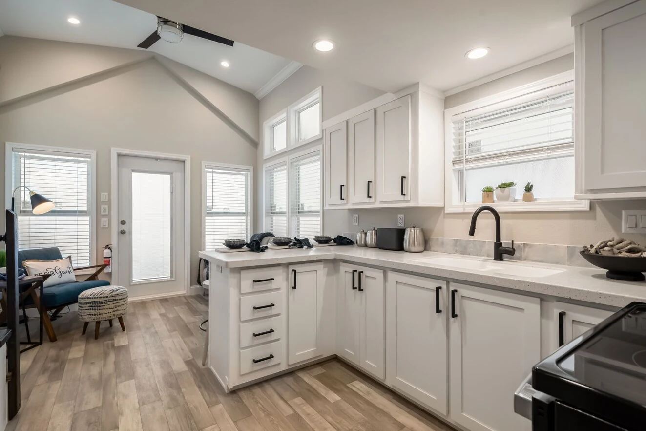 chariot_eagle_eagle_eg12341a_kit_liv_0481_1_1673903426671_1323_12 Modern kitchen with white cabinets, black fixtures, light wood flooring, and stainless steel appliances, adjacent to a small sitting area with natural light from multiple windows.