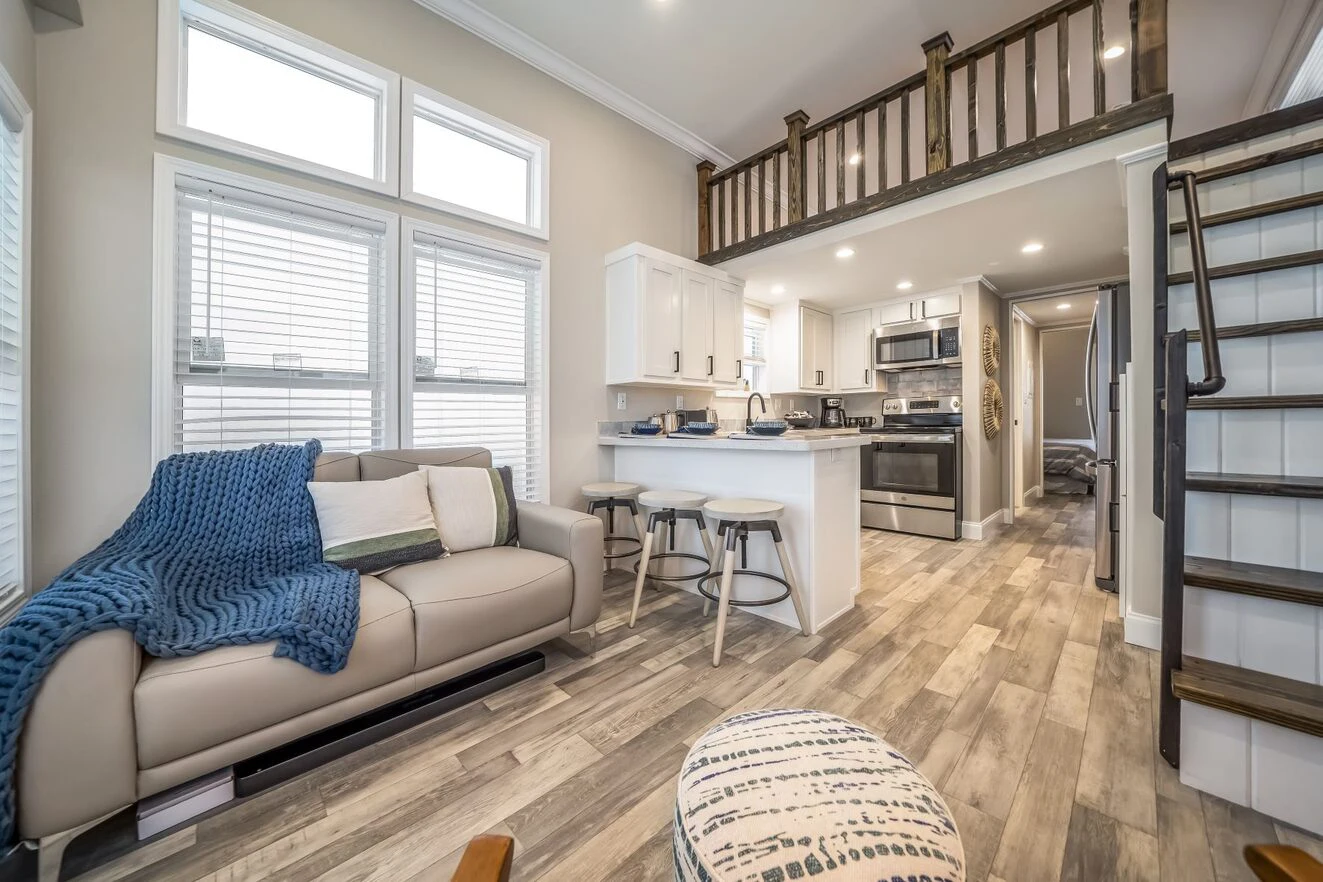 chariot_eagle_eagle_eg12341a_liv_0202_1_1673903420581_1323_12 Modern tiny house interior with a sofa, kitchen island with stools, stainless steel appliances, loft area, and wood staircase. Natural light comes through large windows.